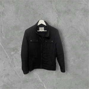 Svart quiltad jacka från Bläck - Svart quiltad pufferjacka från Bläck med snygga detaljer som dragkedja och knappar på bröstfickorna. Jackan har hög krage och två snedställda fickor med dragkedja. Perfekt för dig som gillar stilren och funktionell design.