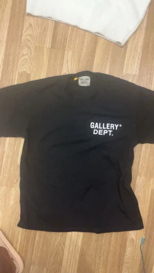 Svart t-shirt från Gallery Dept. - Svart t-shirt från Gallery Dept. med vit logga tryckt på bröstet och stor text på ryggen. Klassisk passform med korta ärmar och rund hals. Tillverkad i mjuk bomull för en skön känsla. Perfekt för dig som gillar streetwear och tydliga prints.