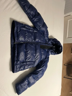 Mörkblå pufferjacka med huva - Säljer en tjock blå dunjacka med mörkblått foder från J Lindeberg med huva och två stora fickor framtill. Perfekt för kalla dagar och har en schysst, lite oversized passform.
