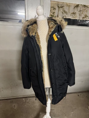 Svart Parajumpers parkajacka med päls - Snygg och varm vinterjacka fràn Parajumpers i svart. Jackan har en stor, fluffig huva och den klassiska gula PJS-remmen vid kragen. Passar perfekt som vardagsjacka under kalla vintermånader. Storlek: XS - passar även S Skick: Mycket bra, knappt använd och inga defekter Köpt för några år sedan men inte använt mer än 2 vintrar !!  En kvalitetsjacka som häller dig varm länge och har mycket kvar att ge. Äkta päls 
