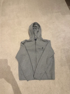 Grå hoodie från Polo Ralph Lauren - Snygg grå hoodie från Polo Ralph Lauren med dragkedja och huva. Klassisk design med Polo-logga broderad på bröstet och två fickor framtill. Perfekt för chill dagar och enkel att matcha med jeans eller joggers. Storlek L i barnstorlek och passar dig mellan 150cm till 160 cm