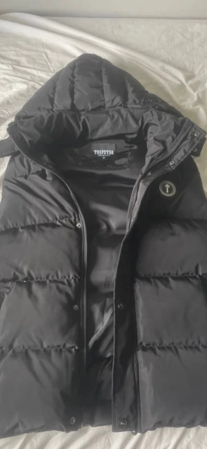 Svart puffervest från Trapstar - Svart puffervest från Trapstar med stor huva och broderad logga på ryggen. Jackan har tryckknappar med logotyp, dragkedja och ett runt Trapstar-märke på bröstet. Materialet är glansig polyester och jackan är vadderad för extra värme. (Om bilderna ser zoomade ut eller skumma så är det för jag screenshotat från mitt konto på vinted)