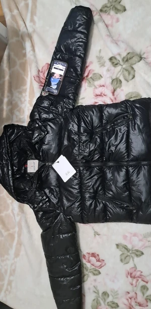 Svart Moncler maya dunjacka med huva - Svart glansig dunjacka från Moncler med huva och dragkedja framtill. Jackan har två snedställda fickor med dragkedja och klassisk Moncler-logga på ärmen . Insidan har ett färgglatt serietryck och märkning för äkthet. Perfekt för kalla dagar. Den har scan och är helt ny oanvänd med etikett 