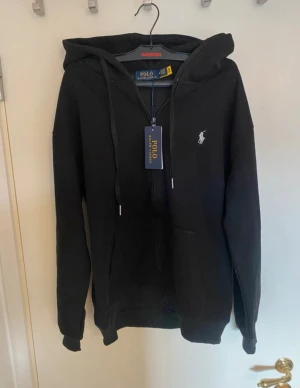 Svart hoodie från Polo Ralph Lauren - Svart hoodie från Polo Ralph Lauren med vit broderad logga på bröstet. Tröjan har huva med snörning, dragkedja framtill och två fickor. Perfekt för chill dagar och enkel att matcha med jeans eller joggers.