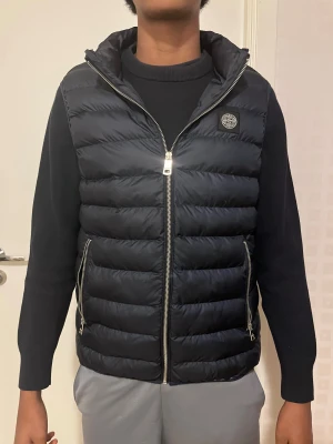 Väst från Stone Island - Snygg svart dunväst från Stone Island. Västen har två sidofickor med dragkedjal. Perfekt att slänga över en hoodie för extra värme och stil. Nytt pris (1899kr)