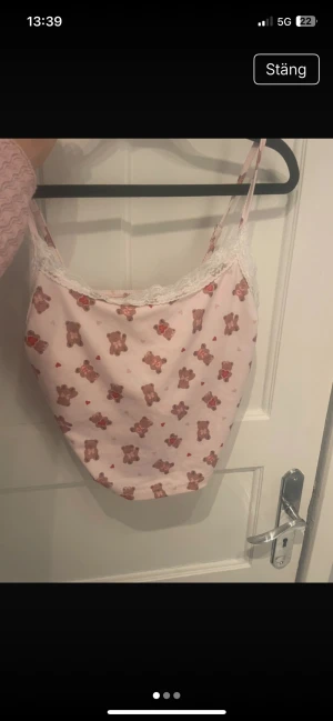 Söt rosa linnetopp med nallemönster - Rosa linnetopp från SHEIN med gulligt nallemönster och små röda hjärtan. Toppen har justerbara smala axelband, spetsdetaljer längs halsringningen och är croppad i modellen. Materialet är mjukt och lätt polyester med lite stretch för skön passform.