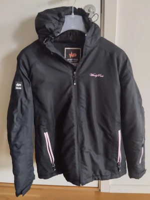 Svart vindjacka från Windy Peak - Svart vindjacka från Windy Peak med huva och rosa detaljer vid dragkedjorna. Jackan har lång ärm, flera fickor med dragkedja och sportig design. Perfekt för dig som gillar funktionella och snygga ytterplagg. Snygg logga på bröstet och ärmen.