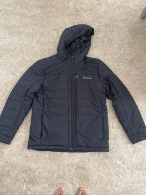 Svart pufferjacka Peak Performance - Svart pufferjacka från Peak Performance med huva och hel dragkedja. Jackan har diskret logga på bröstet, quiltad design och praktiska fickor. Perfekt för kalla dagar och enkel att matcha med allt. Materialet är syntet och jackan är långärmad.