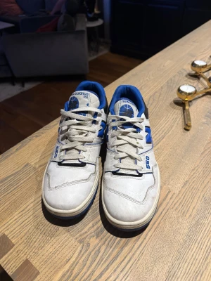 New Balance 550 sneakers vit/blå - Säljer ett par New Balance 550 sneakers i vit och blå färg med svarta detaljer. Skorna har snörning, rund tå och platt sula. Ovandelen är i skinn och syntet med blå meshfoder. Ikonisk N-logga på sidan och tydlig 550-märkning. Perfekt för dig som gillar retro basketstil.