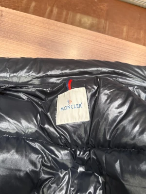 Svart Moncler dunjacka med röd/vit detalj - Snygg svart dunjacka från Moncler med glansig finish och klassisk pufferstil. Jackan har dragkedja och coola röd/vita detaljer längs nederkanten. Perfekt för kalla dagar när du vill hålla stilen och värmen.