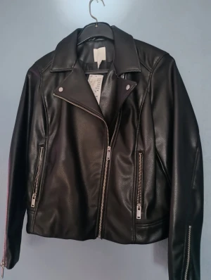 Svart skinnjacka med dragkedjor - Snygg svart skinnjacka med klassisk bikerkrage och silvriga dragkedjor på fickor och ärmslut. Jackan har en rak passform och coola detaljer som ger en edgy vibe. Perfekt för dig som vill ha en stilren och tidlös look. Knappt använd, storlek L 