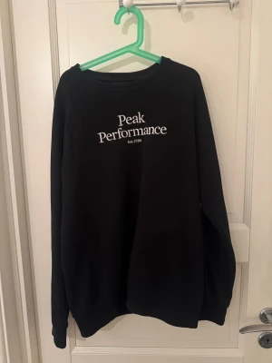 Svart Peak Performance sweatshirt - Svart sweatshirt från Peak Performance med vit broderad logga på bröstet. Tröjan har rund hals, ribbade muddar och lång ärm. Perfekt för chill dagar och enkel att matcha med jeans eller mjukisbyxor.