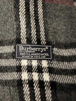 Rutig grå halsduk från Burberry - Snygg halsduk från Burberry i klassiskt rutigt mönster med grå bas, vita och svarta linjer samt röda detaljer. Gjord av 100% lammull, mjuk och varm med fransar i ändarna. Perfekt för att lyfta din vinterstil med en ikonisk London-vibe.