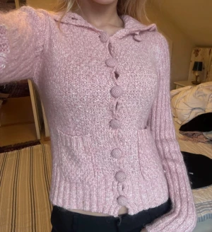 Ljusrosa stickad kofta med stora knappar - Supermysig ljusrosa stickad kofta med stora dekorativa knappar framtill och två fickor. Koftan har långärmade ribbade ärmar och en söt krage med knappdetaljer. Perfekt för dig som gillar gulliga och unika plagg med lite retrovibe.