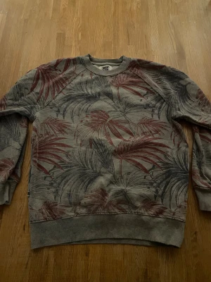 Grå sweatshirt med palmtryck Sweet SKTBS - Grå sweatshirt från Sweet SKTBS i storlek XS med coolt palmbladsmönster i mörkrött och mörkgrått. Tröjan har rund hals, ribbade muddar och långärmad passform. Perfekt för dig som gillar streetwear och snygga prints.