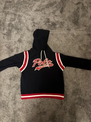Mörkblå hoodie Polo Ralph Lauren - Snygg mörkblå hoodie från Polo Ralph Lauren med röd och vit logga framtill. Tröjan har huva med snörning, långa ärmar och coola ränder i rött och vitt på axlar, ärmslut och nederkant. Perfekt för dig som gillar sportig och street-inspirerad stil.
