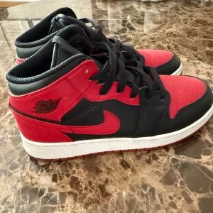 Nike Air Jordan 1 Mid Svart/Röd - Säljer ett par Nike Air Jordan 1 Mid i klassisk svart och röd färg med vita sulor. Skorna har snörning, rund tå och är tillverkade i skinn och syntet. Perfekta för dig som vill sticka ut med en ikonisk sneaker.