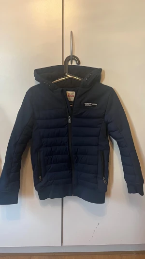 Blå quiltad jacka från Vingino - Snygg mörkblå pufferjacka från Vingino med huva och dragkedja framtill. Jackan har quiltade partier, två fickor med dragkedja och ribbade muddar. Perfekt för kyliga dagar och har en sportig vibe.