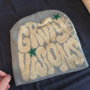 Grå mössa med tryck och stjärnor - Grå stickad mössa med stort beige tryck 'GROOVY VISIONS' framtill, gröna stjärnor och ett coolt spindelnätsmönster på sidan. Mjuk och skön, perfekt för att sticka ut i mängden under kalla dagar. Hej jag vill sälja min mössa helt ny jag vill ha pengar de darför jag säljer min mössa