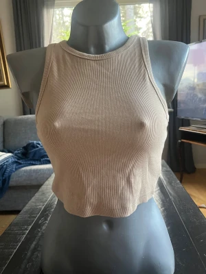 Beige ribbad croppad linnetopp - Säljer en beige croppad linnetopp i ribbad bomull. Toppen är ärmlös med rund halsringning och har en enkel, stilren look som passar perfekt till jeans eller kjol. Perfekt för varma dagar eller lager på lager.