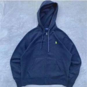 Mörkblå hoodie från Polo Ralph Lauren - Snygg mörkblå hoodie från Polo Ralph Lauren med dragkedja och huva. Tröjan har snörning vid halsen, klassisk logga i gult på bröstet och stora fickor framtill. Perfekt för chill dagar och enkel att matcha med jeans eller joggers. Står M i men den har krympt i tvätten så sitter mer som xs/s