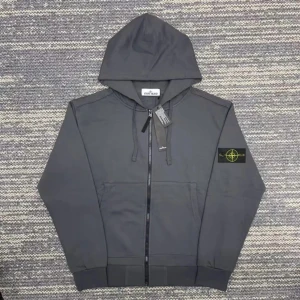 Mörkgrå hoodie från Stone Island - Säljer en mörkgrå hoodie från Stone Island med huva och dragkedja framtill. Tröjan har en klassisk patch med logga på vänster ärm, snörning vid huvan och två stora fickor fram. Perfekt för dig som gillar streetwear och snygga detaljer.