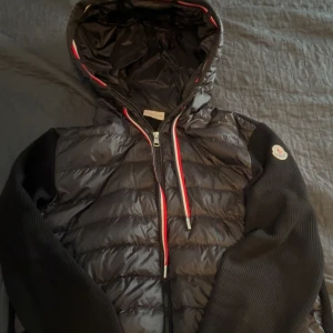 Svart Moncler pufferjacka med huva ÄKTA - Snygg svart pufferjacka från Moncler med quiltad front, stickade ärmar och huva. Jackan har dragkedja och röda/vita detaljer på snören och huvkant.   Kan gå ner i pris vid en snabb affär