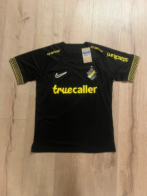 AIK svart matchtröja Nike 2024 - AIK:s officiella matchtröja från Nike, svart med gula detaljer och tryck. Kortärmad med gulrandiga muddar, klubbmärke på bröstet och sponsorer som Truecaller och SVEA. Tillverkad i lätt, ventilerande Dri-FIT-material som passar perfekt för fotbollsträning.