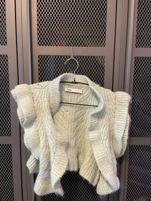 Stickad ljusgrön väst från Zara - Superfin stickad väst från Zara i en ljusgrön ton. Västen har breda volangdetaljer längs axlarna och ett snyggt kabelstickat mönster. Den är croppad och har en mjuk, fluffig känsla som gör den extra mysig. Perfekt att styla över en långärmad tröja.