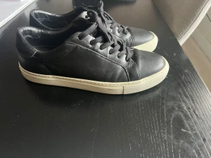 Svarta sneakers från Rizzo i skinn - Stilrena svarta sneakers från Rizzo i mjukt skinn med vita, platta sulor. Klassisk design med rund tå och svarta snören. Perfekta för en clean och enkel look. Passar till det mesta och är superenkla att matcha.