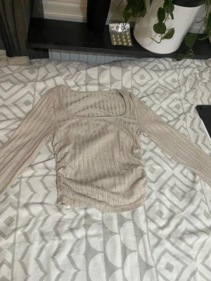 Beige ribbad långärmad topp - Säljer en beige ribbad topp med lång ärm och fyrkantig halsringning. Toppen har snygg snörning på sidan som ger en cool rynkad effekt. Perfekt att styla med jeans eller kjol för en chill look.