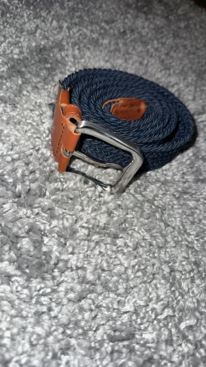 Mörkblått flätat bälte med skinn 105cm - Snyggt mörkblått flätat bälte med detaljer i brunt skinn och ett klassiskt silverfärgat spänne. Bältet har en stilren look och passar perfekt till jeans eller chinos. Flätad design gör det enkelt att justera längden efter behov.