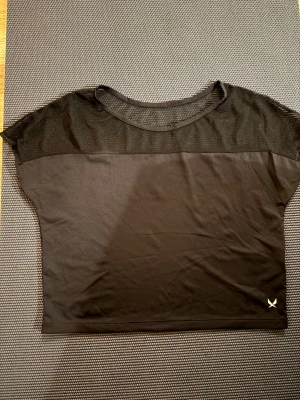 Svart mesh topp från Soc - Svart croppad topp från ICANIWILL med meshparti över axlar och bröst. Rund halsringning och korta ärmar. Materialet är mjukt och stretchigt, perfekt för träning. Liten vit logga nere till höger. Snygg och sportig vibe!
