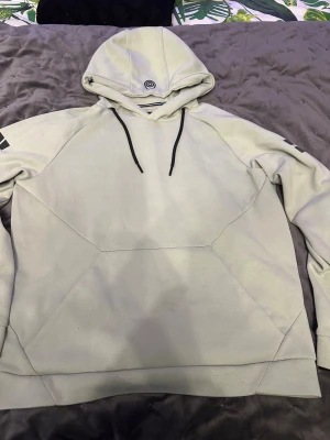 Ljus sage grön hoodie från Sail Racing S - Snygg ljus sage grön hoodie från Sail Racing i storlek S. Tröjan har svart tryck på huvan och båda ärmarna, samt svarta snören vid huvan. Tillverkad i mjuk bomull med coola geometriska sömmar framtill. Perfekt för dig som gillar stilrena och sportiga plagg.