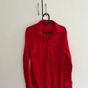 Röd plisserad skjorta med knappar - Röd längre skjorta med plisserad struktur och klassisk krage. Skjortan har långa ärmar och knäppning framtill. Uppskattningsvis storlek S. 100kr+frakt