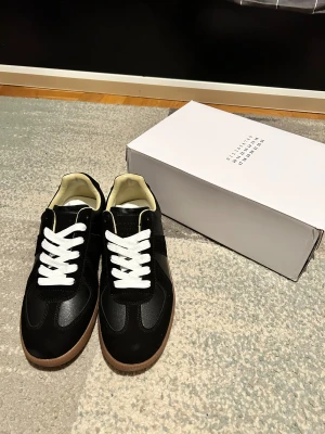 Maison Margiela Gats  - Maison Margiela sneakers i svart skinn och mocka med vita snören och beige innersida. Snygg gummisula i brun ton med unikt mönster undertill. Klassisk rund tå och platt sula, stilren design med diskreta detaljer. Kommer med originalkartong.