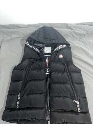 (Pris kan diskuteras) Svart dunväst från Moncler med huva  - Svart dunväst från Moncler med stor logga på insidan och broderad Moncler-patch på bröstet. Västen har huva med Moncler-tryck längs kanten, två sidofickor med dragkedja och quiltad design. Insidan har en cool serietidningsetikett och blanka materialdetaljer.