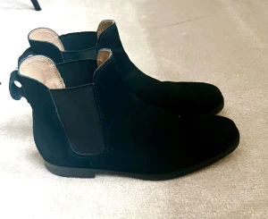 Svarta Chelsea boots från Ralph Lauren Polo Ralph Lauren - Svarta Chelsea boots i mocka från Polo Ralph Lauren. Snygg, klassisk siluett med elastiska paneler på sidorna och platt sula. Insidan är ljusbeige och har en rund tå. Perfekta för dig som gillar stilrena och tidlösa skor.