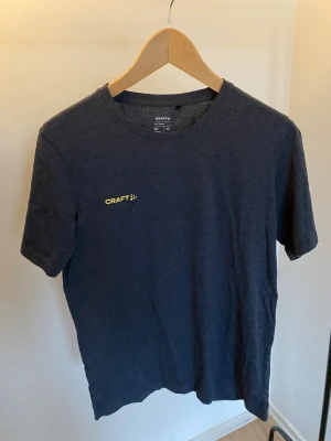 Mörkgrå Craft Sweden Collection t-shirt - Snygg mörkgrå t-shirt från Craft Sweden Collection med gul logga fram och stort SWEDEN-tryck på ryggen. Klassisk rund hals och korta ärmar. Perfekt för dig som gillar sportig stil och vill visa lite svensk stolthet. Materialet är mjukt och skönt bomull.