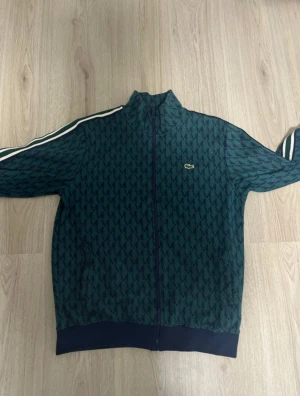 Gröna mönstrade mjukisbyxor från Lacoste - Säljer ett par gröna mjukisbyxor från Lacoste med mörkblå diamantmönster, vita ränder längs sidorna och snörning i midjan. Byxorna har en relaxed passform och är perfekta för chill dagar. Materialet är mjukt och skönt, med klassisk Lacoste-logga på benet.