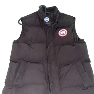 Svart dunväst från Canada Goose - Svart dunväst från Canada Goose med klassisk logga på bröstet. Västen har hög krage, två stora fickor framtill och stängs med både dragkedja och knappar. Perfekt för lager-på-lager och håller dig varm under kyliga dagar.