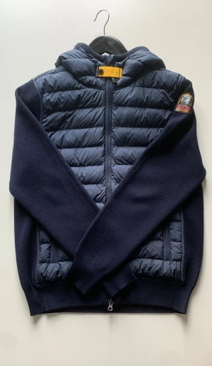 Parajumpers Illuga Cardigan - Säljer en snygg mörkblå cardigan från Parajumpers i storlek XS. Jackan har quiltad framsida och huva, stickade ärmar och rygg samt dragkedja framtill. Klassisk patch på ärmen och gul detalj vid halsen. Perfekt för de lite varmare vinterdagarna. Cardiganen är i nyskick, bara använd ett fåtal gånger. Allt medföljer såsom box, brev och tags. Pris kan diskuteras. Kontakta vid intresse.