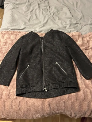 Svart jacka från Rut&Circle, strl 36 - Snygg svart jacka från Rut&Circle i storlek 36. Jackan har en stilren look med struktur i tyget, silverfärgade dragkedjor framtill och på fickorna. Rak passform och lång ärm, perfekt för dig som gillar en clean och modern stil.