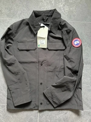 Canada Goose jacka - Jacka från Canada Goose med klassisk krage, två bröstfickor med lock och knäppning framtill. Jackan har det ikoniska Canada Goose-märket på vänster ärm och är tillverkad i slitstark polyester med slät insida. Använd två gånger 