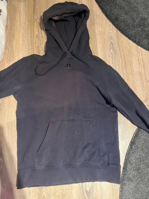 Svart hoodie från J.Lindeberg  - Svart hoodie från J.Lindeberg med broderat logotyp på bröstet och palmer samt texten 'Beach Tour Spring Summer 2019' på ryggen. Tröjan har huva med snörning, känguruficka och är tillverkad i mjuk bomull. Perfekt för chill dagar och streetstyle.