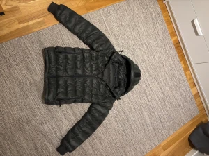 Svart pufferjacka Peak Performance strl. xs/170 - Svart pufferjacka från Peak Performance med huva och dragkedja framtill. Jackan har quiltad design, två sidofickor med dragkedja och ribbade muddar vid ärmslut. Perfekt för kalla dagar och har en clean, sportig look.