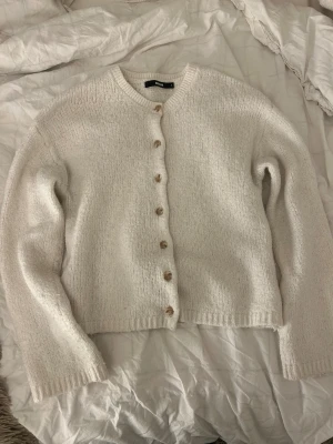 Vit stickad kofta  - Mysig vit stickad kofta från Bik Bok i storlek S. Koftan har rund hals, långa ärmar och snygga beigea knappar framtill. Perfekt att slänga på sig när det är lite kyligt ute. Ribbstickade muddar vid ärmslut och nederkant ger en chill vibe.