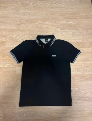 Svart slim fit piké från BOSS - Svart pikétröja från BOSS med vit logga på bröstet och vita ränder på krage och ärmslut. Slim fit-modell med stretch för extra komfort. Klassisk pikékrage och korta ärmar, perfekt för en clean och stilren look.