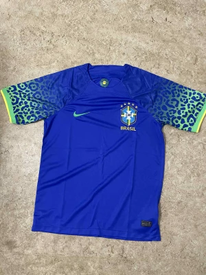Blå Brasil fotbollströja Nike - Säljer en blå Brasil fotbollströja från Nike med korta ärmar. Ärmarna har leopardmönster i grönt och blått med gula detaljer vid mudden. Tröjan har Brasil-logga och Nike swoosh på bröstet. Materialet är lätt och andas, perfekt för match eller träning.