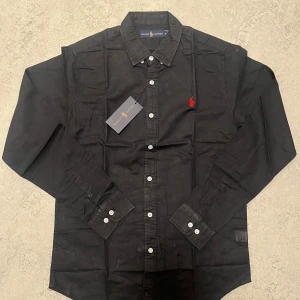 Svart skjorta från Ralph Lauren S - Svart skjorta från Ralph Lauren med klassisk krage och vita knappar framtill. På bröstet finns den ikoniska röda Polo-loggan broderad. Skjortan är tillverkad i bomull och har lång ärm. Perfekt för dig som gillar stilrena och tidlösa plagg. Byten funkar.